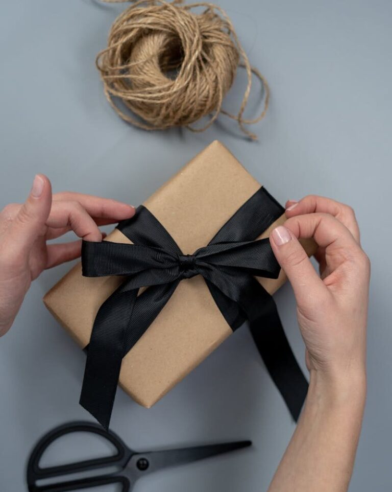 Hands tying a black ribbon on a craft paper gift box, next to scissors and twine.