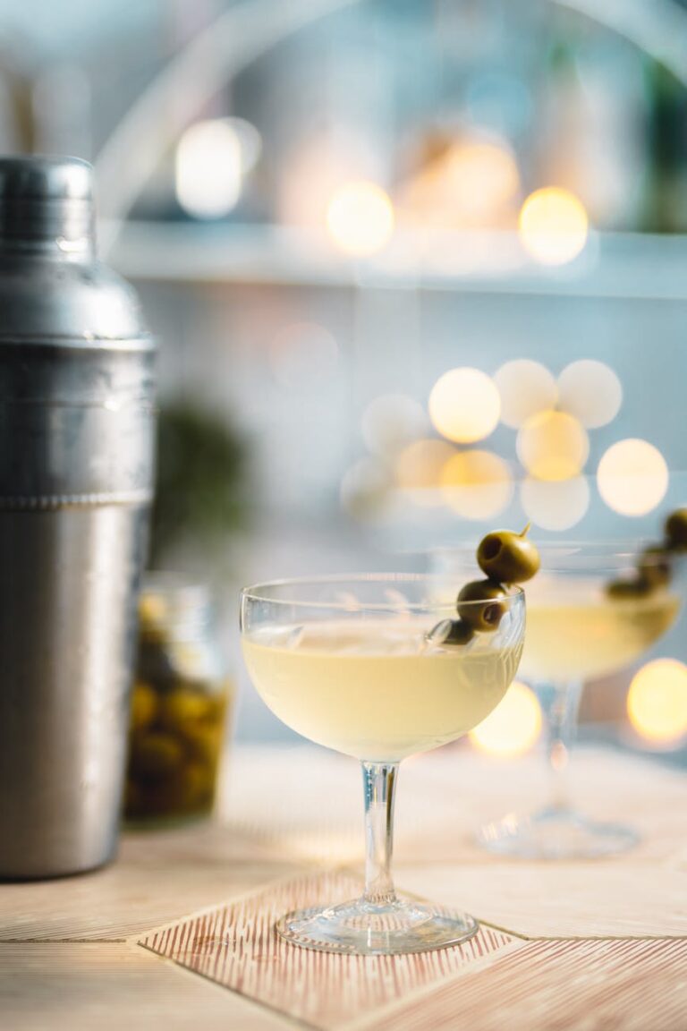 A stylish martini garnished with olives, alongside a cocktail shaker on a blurred bokeh background.
