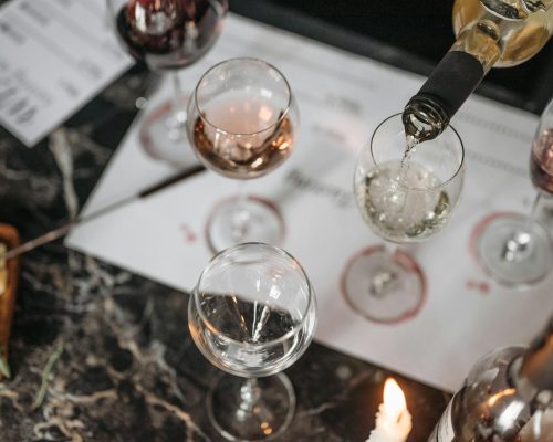 Overhead view of a wine tasting setup on a marble table with white wine being poured and cheese platter nearby.